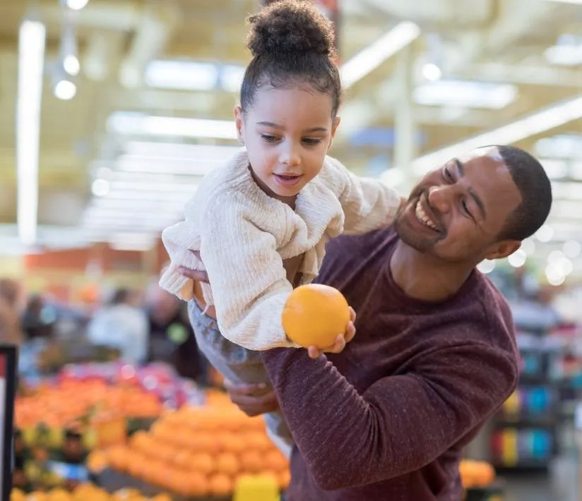 Man holding up his daughter who is holding an orange in grocery story