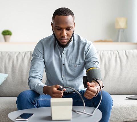man with ongoing healthcare needs sitting on the couch and taking his blood pressure using a blood pressure machine