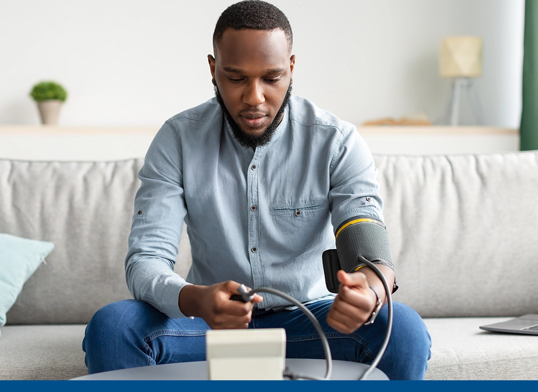 man with ongoing healthcare needs sitting on the couch and taking his blood pressure using a blood pressure machine