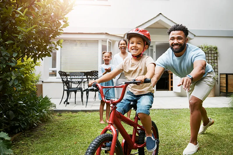 Proud dad teaching his young son to ride bicycle while wearing a helmet for safety in their family home garden, mom and daughter celebrate in background.