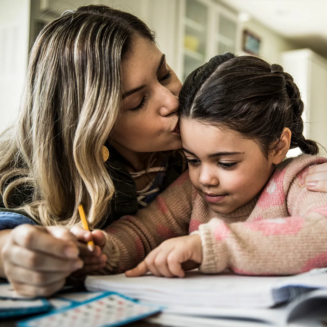 Madre ayudando a su hija pequeña con la tarea y besándola en la frente