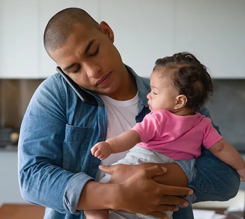 Padre joven sosteniendo a su bebé en brazos mientras habla por teléfono con un agente dedicado sobre la inscripción en un plan de salud de Florida Blue para 2026
