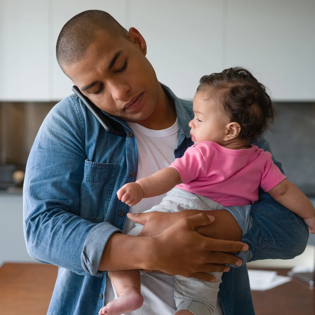 Padre joven sosteniendo a su bebé en brazos mientras habla por teléfono con un agente dedicado sobre la inscripción en un plan de salud de Florida Blue para 2026