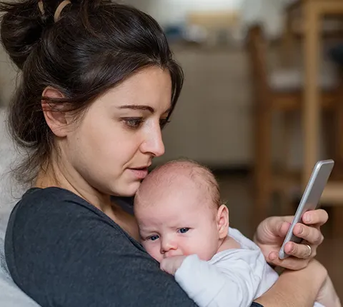 Joven madre sosteniendo a su bebé en brazos mientras chatea en vivo en su teléfono móvil con un agente dedicado sobre la inscripción en un plan de salud de Florida Blue para 2026