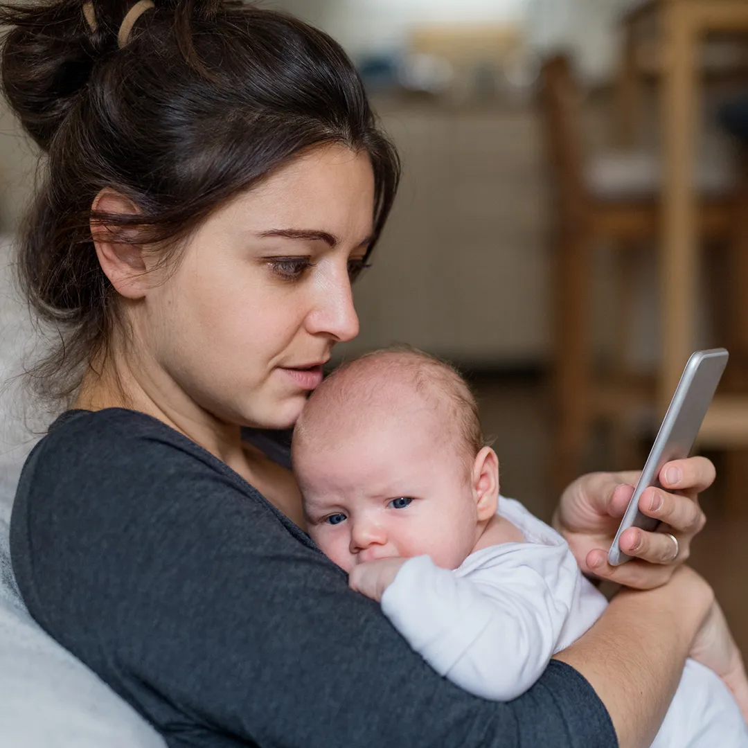 Joven madre sosteniendo a su bebé en brazos mientras chatea en vivo en su teléfono móvil con un agente dedicado sobre la inscripción en un plan de salud de Florida Blue para 2026